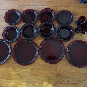 May include: A collection of ruby red glassware, including plates, bowls, and small glasses. The plates and bowls have a decorative, scalloped edge. The glasses are small and have a unique shape. The set is arranged on a wooden surface.