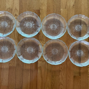 May include: Eight clear glass plates with a floral design in the center and a scalloped edge. The plates are arranged in two rows of four on a wooden surface.