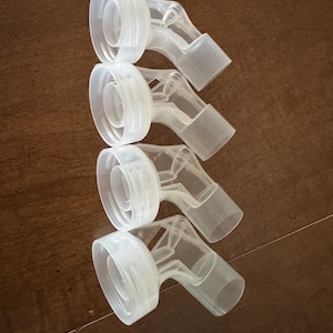 May include: Four clear plastic breast pump connectors are arranged in a row. Each connector has a wide, circular opening on one end, a curved midsection, and a rectangular spout on the other end. The connectors are made of translucent plastic.