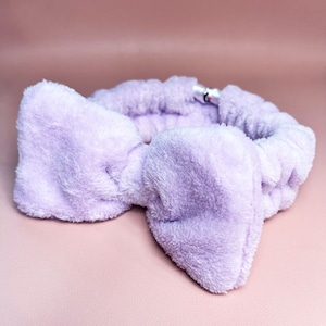 May include: A purple fluffy headband with a bow. The headband is made of soft, plush material and is designed to hold hair back while applying makeup or skincare.