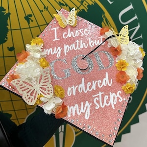 May include: A pink glitter graduation cap decorated with white flowers, orange and yellow accents, and butterfly cutouts. The cap features the text "I chose my path but GOD ordered my steps" in white and silver.