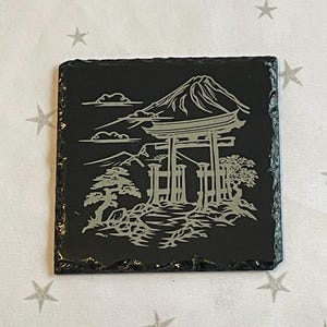 Può includere: Sottobicchiere in ardesia nera con un designo inciso in argento del Monte Fuji, un torii e alberi.