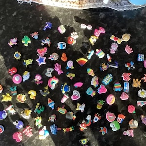May include: A collection of colourful shoe charms featuring various designs, including animals, food, and emojis. The charms are made of rubber and have a variety of colours, including pink, blue, green, yellow, and red.