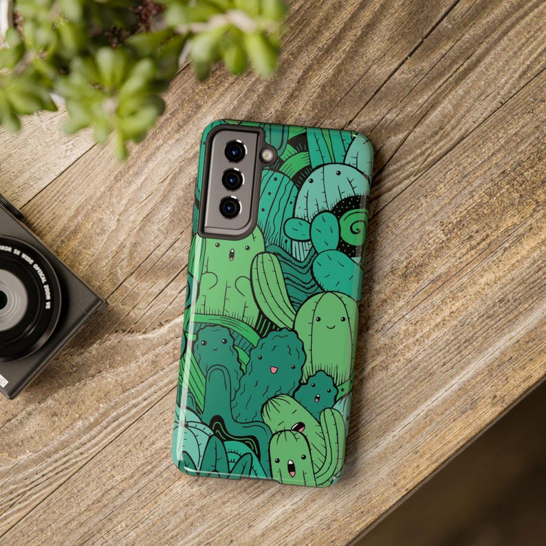 Peut inclure: Une coque de t&eacute;l&eacute;phone verte avec un design fantaisiste repr&eacute;sentant plusieurs cactus avec des visages souriants. La coque a une d&eacute;coupe pour l'appareil photo avec trois objectifs.
