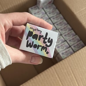 Puede incluir: Una pequeña caja blanca con el texto "mystery party worm" en negro y colores del arcoíris se sostiene en una mano. La caja está frente a una caja de cartón más grande llena de cajas similares. Las cajas parecen ser un regalo de fiesta.