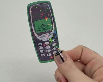 May include: A sticker of a retro cell phone with a green border and a purple screen. The screen displays the text "Sugar Crash Club" in white. The sticker has a holographic effect with colorful star accents.