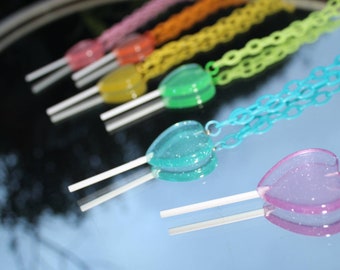 May include: A rainbow assortment of heart-shaped lollipops on sticks with glitter accents. Each lollipop is attached to a colorful chain necklace.