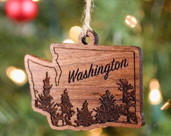Può includere: Un ornamento natalizio in legno a forma dello stato di Washington con la parola "Washington" incisa su di esso. L'ornamento presenta una silhouette di alberi all'interno del contorno dello stato.