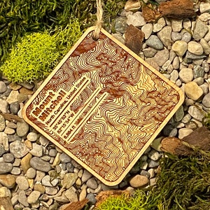 May include: Wooden Mount Mitchell ornament with a topographic map design. The ornament is square with rounded corners and includes the mountain's coordinates. It hangs from a natural twine string. The background features moss, small stones, and dried plant material.