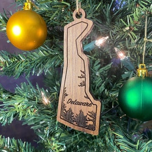 May include: Wooden Christmas ornament shaped like the state of Delaware with the word "Delaware" engraved on it. The ornament is hanging on a green Christmas tree.