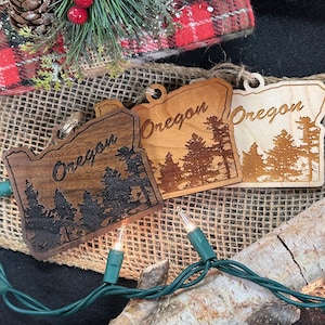 May include: Two wooden ornaments shaped like the state of Oregon, with the word "Oregon" engraved on each. The ornaments are on a burlap background with Christmas lights.