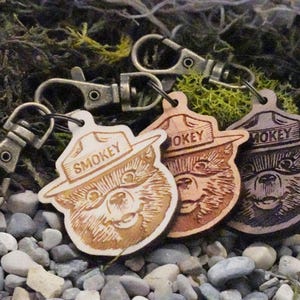 May include: Three wooden keychains featuring Smokey Bear, the mascot of the U.S. Forest Service. Each keychain has a different wood stain color: light brown, dark brown, and black. The keychains are attached to silver key rings.