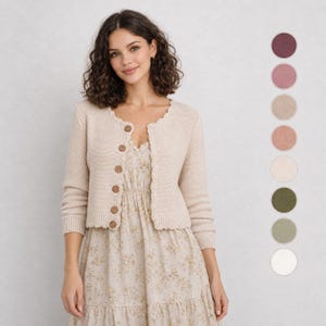 May include: A beige knit cardigan with a scalloped edge and brown buttons, worn over a cream-coloured dress with a small floral pattern. A vertical column of nine circular colour swatches is displayed to the right of the garment.