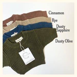 May include: Four long-sleeve knit sweaters stacked vertically in colors labeled as Cinnamon, Rye, Dusty Sapphire, and Dusty Olive. Each sweater features a ribbed crew neckline and a white brand tag reading Olive & Cradle attached by a string.