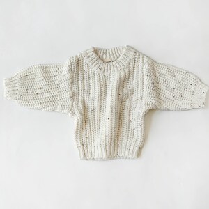 May include: A cream-colored chunky knit sweater featuring a ribbed texture with subtle brown and black flecks throughout. The garment has a round neckline, long sleeves with ribbed cuffs, and a ribbed hemline, showcasing a thick, textured yarn construction.