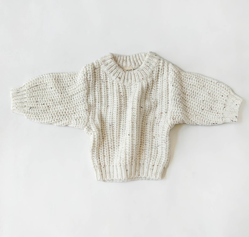 May include: A cream-colored chunky knit sweater featuring a ribbed texture with subtle brown and black flecks throughout. The garment has a round neckline, long sleeves with ribbed cuffs, and a ribbed hemline, showcasing a thick, textured yarn construction.