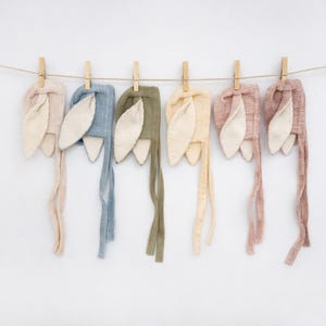 May include: Six pastel-colored baby bonnets with bunny ears hanging from a clothesline. The bonnets are in shades of pink, blue, green, and yellow, with contrasting cream-colored ears. Each bonnet has long ties.