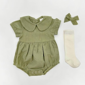 May include: A green linen baby romper with a large peter pan collar and short sleeves. The romper has a gathered skirt and snaps at the bottom. The set also includes a pair of white knit socks and a green bow headband.