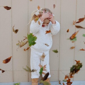 May include: A young girl wearing a white knitted sweater and matching pants stands in front of a beige wall. She is smiling and has her hands on her head. Autumn leaves are falling around her.