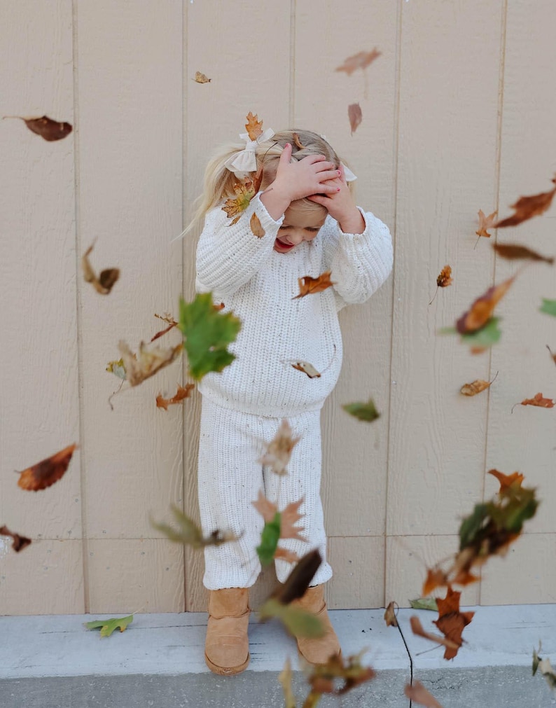 May include: A young girl wearing a white knitted sweater and matching pants stands in front of a beige wall. She is smiling and has her hands on her head. Autumn leaves are falling around her.
