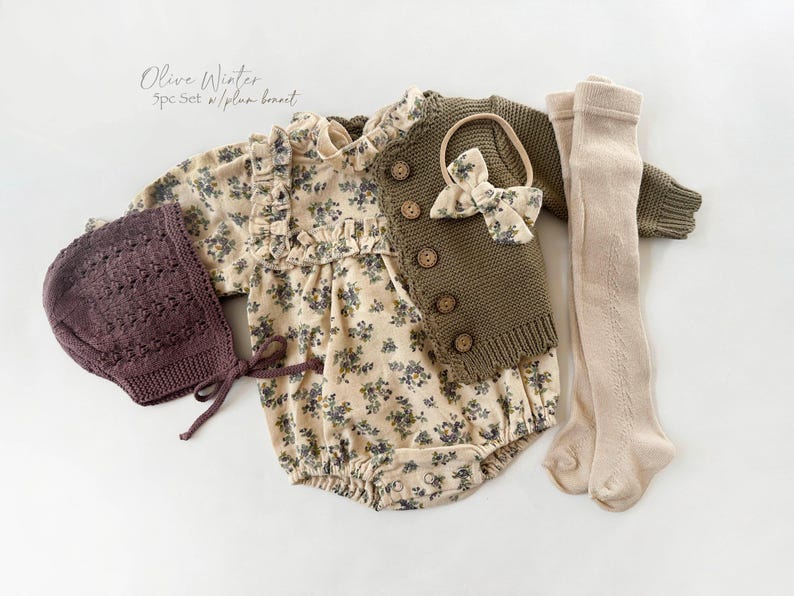 May include: A baby outfit set with a floral print bodysuit, a brown knitted cardigan, a brown knitted hat, and beige tights. The set also includes a brown bow headband.