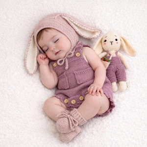 May include: A sleeping baby in a mauve knitted bunny hat and overalls, with matching booties. The outfit has button details. A crocheted bunny doll with a carrot in its pocket lies next to the baby on a white surface.
