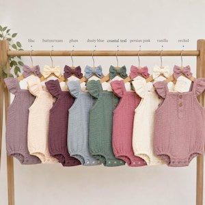 Puede incluir: Una colección de peleles para bebés en varios colores pastel, incluyendo lila, crema de mantequilla, ciruela, azul polvoriento, verde azulado costero, rosa persa, vainilla y orquídea. Cada pelele tiene un lazo a juego y se muestra en una percha de madera.
