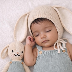 Puede incluir: Un bebé dormido con un gorro de conejito color crema con orejas largas y un pelele de punto azul claro. Una muñeca de conejito a juego con una cara sonriente y detalles de botones descansa junto al bebé. La escena se desarrolla sobre una superficie blanca y suave.