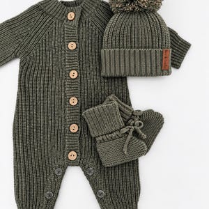 Peut inclure: Une barboteuse en tricot côtelé vert olive avec six boutons en bois clair sur le devant, des chaussons assortis avec lacets et un bonnet côtelé orné d'un gros pompon et d'une petite étiquette marron marquée Fox & Cradle.