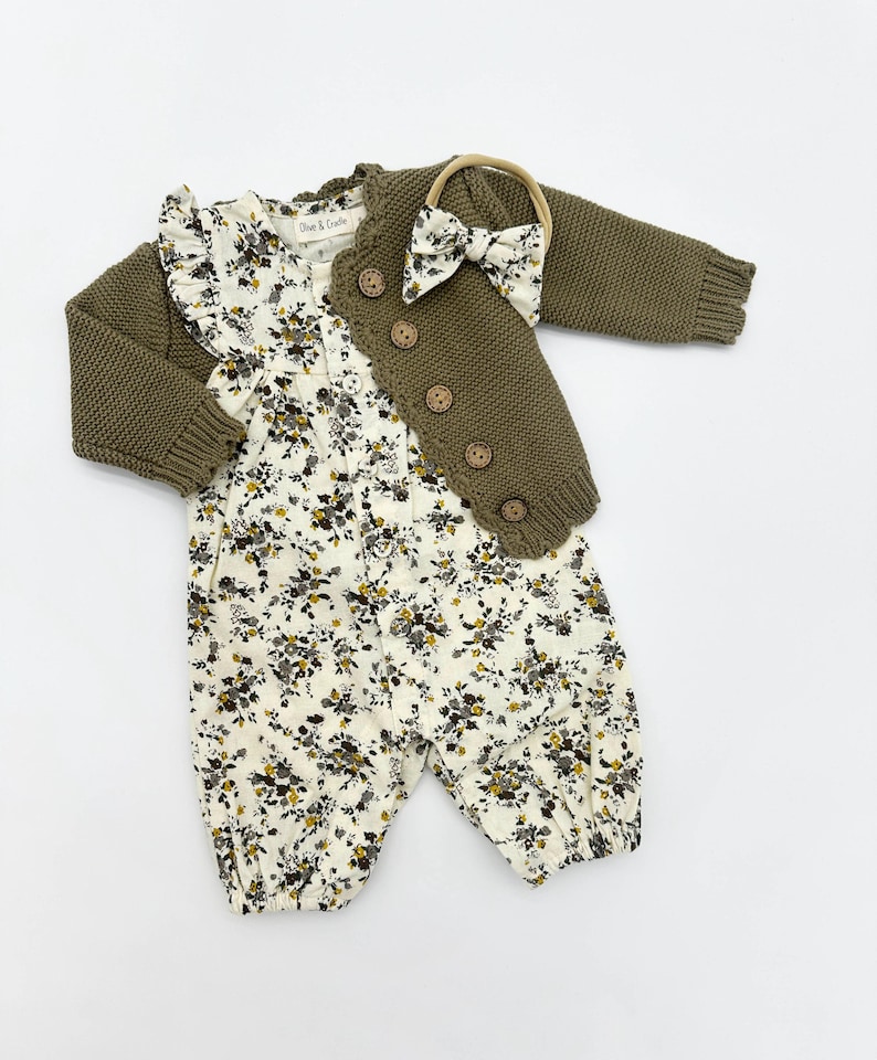 May include: A baby outfit set featuring a floral print jumpsuit, a green knitted cardigan with wooden buttons, and a brown headband with a bow.