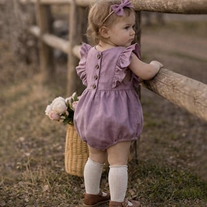 Op de afbeelding: Een kind in een lavendelkleurige romper met ruchesmouwen en houten knopen. Het kind draagt een bijpassende strik, witte kniekousen en bruine leren schoenen. Een rieten mand met bloemen is zichtbaar.