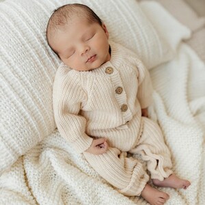 May include: A newborn baby wearing a cream-colored knitted sweater and pants, sleeping on a white knitted blanket. The baby has dark hair and is wearing a cream-colored knitted outfit.