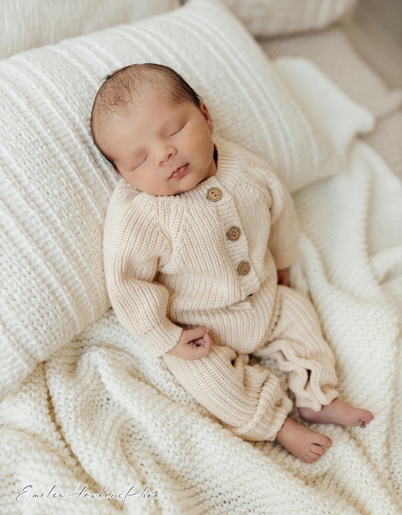 May include: A newborn baby wearing a cream-colored knitted sweater and pants, sleeping on a white knitted blanket. The baby has dark hair and is wearing a cream-colored knitted outfit.