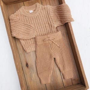 May include: A tan knitted sweater and matching pants set for a baby. The sweater has a crew neck and long sleeves. The pants have a drawstring waistband and are cuffed at the ankles.