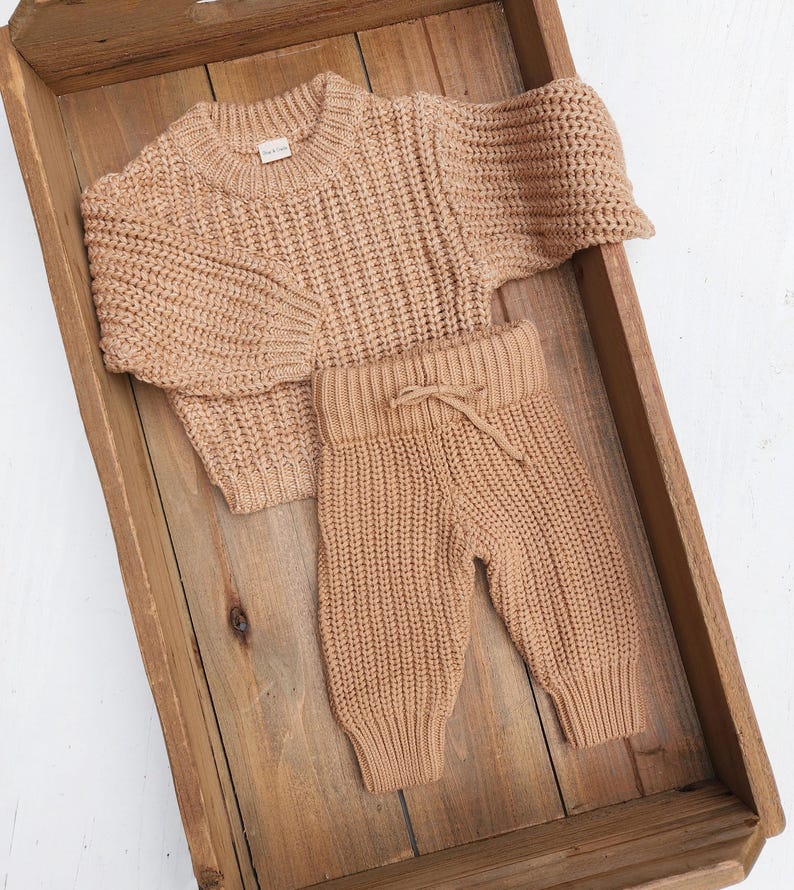 May include: A tan knitted sweater and matching pants set for a baby. The sweater has a crew neck and long sleeves. The pants have a drawstring waistband and are cuffed at the ankles.