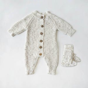 May include: A white knitted baby onesie with brown buttons and matching knitted booties.