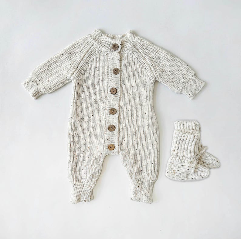 May include: A white knitted baby onesie with brown buttons and matching knitted booties.