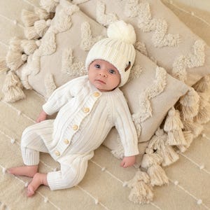 May include: A baby wearing a white knitted hat with a pom pom and a white knitted onesie. The baby is lying on a cream-colored blanket with tassels.