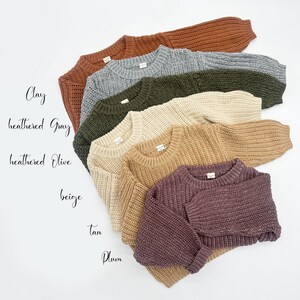 May include: A collection of six long-sleeved knit sweaters in various colors, including clay, heathered gray, heathered olive, beige, tan, and plum. Each sweater features a ribbed crew neckline, ribbed cuffs, and a chunky, textured knit pattern.