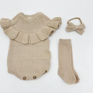 May include: A beige knitted baby outfit with a ruffled neckline, matching socks, and a headband with a bow.