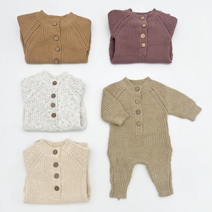 May include: Four knitted baby onesies in different colors and patterns. The onesies have a button closure down the front and long sleeves. The colors are brown, beige, white with brown speckles, and purple.