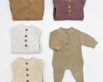 May include: Four knitted baby onesies in different colors and patterns. The onesies have a button closure down the front and long sleeves. The colors are brown, beige, white with brown speckles, and purple.