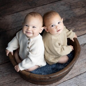May include: Two babies wearing cream and beige knitted sweaters sit in a wooden bowl. The babies are looking at the camera and smiling.