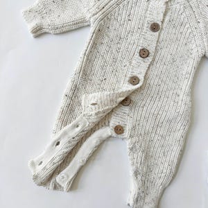 May include: A white knitted baby onesie with brown buttons. The onesie has a speckled texture and long sleeves.