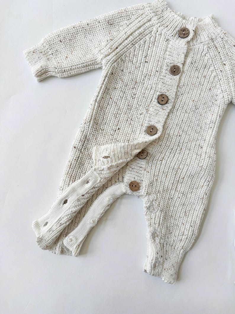 May include: A white knitted baby onesie with brown buttons. The onesie has a speckled texture and long sleeves.