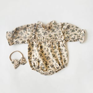 May include: A beige baby onesie with a floral print and ruffled trim. The onesie has long sleeves and a snap closure at the bottom. A matching headband with a bow is included.