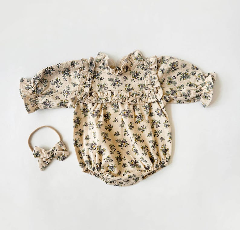 May include: A beige baby onesie with a floral print and ruffled trim. The onesie has long sleeves and a snap closure at the bottom. A matching headband with a bow is included.
