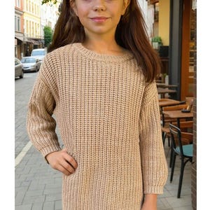 Könnte beinhalten: Ein beigefarbener Langarmpullover mit gerippter Strickstruktur. Der Pullover hat einen Rundhalsausschnitt und eine lockere Passform. Die Person trägt schwarze Leggings. Der Hintergrund zeigt eine europäische Straßenszene mit Gebäuden und einem Café.