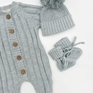 May include: A light blue knitted baby outfit set with a long-sleeved onesie, a beanie with a pom-pom, and booties.