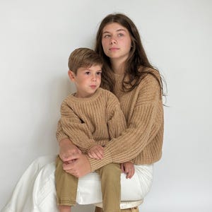 May include: Two matching tan long-sleeve sweaters with a thick ribbed knit texture and crew necklines. The sweaters feature a chunky weave pattern, long sleeves with ribbed cuffs, and a relaxed fit, providing a uniform look for two individuals.
